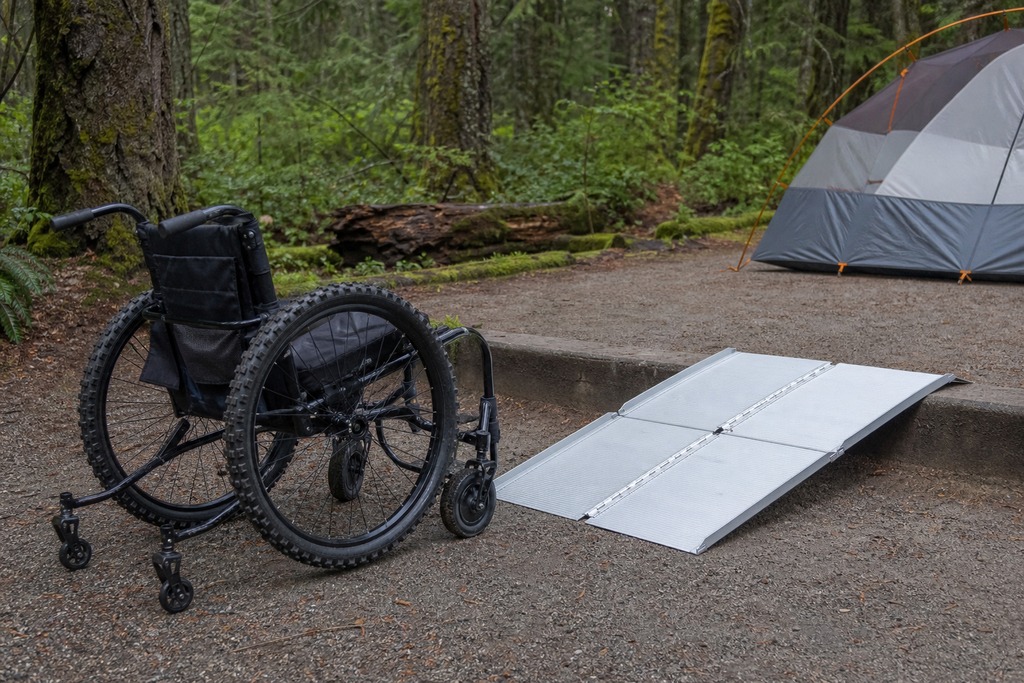 A wheelchair next to a portable ramp leading to a camping area with a tent in a forest setting.