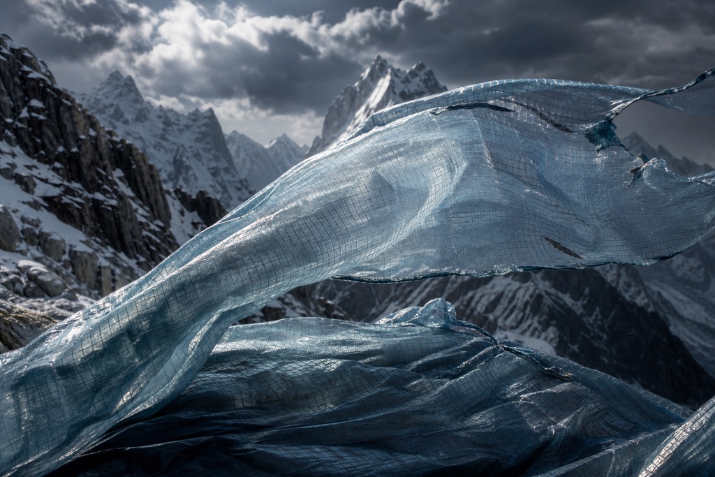 A translucent, textured blue material resembling a graphene sleeping bag billows against a mountainous backdrop.