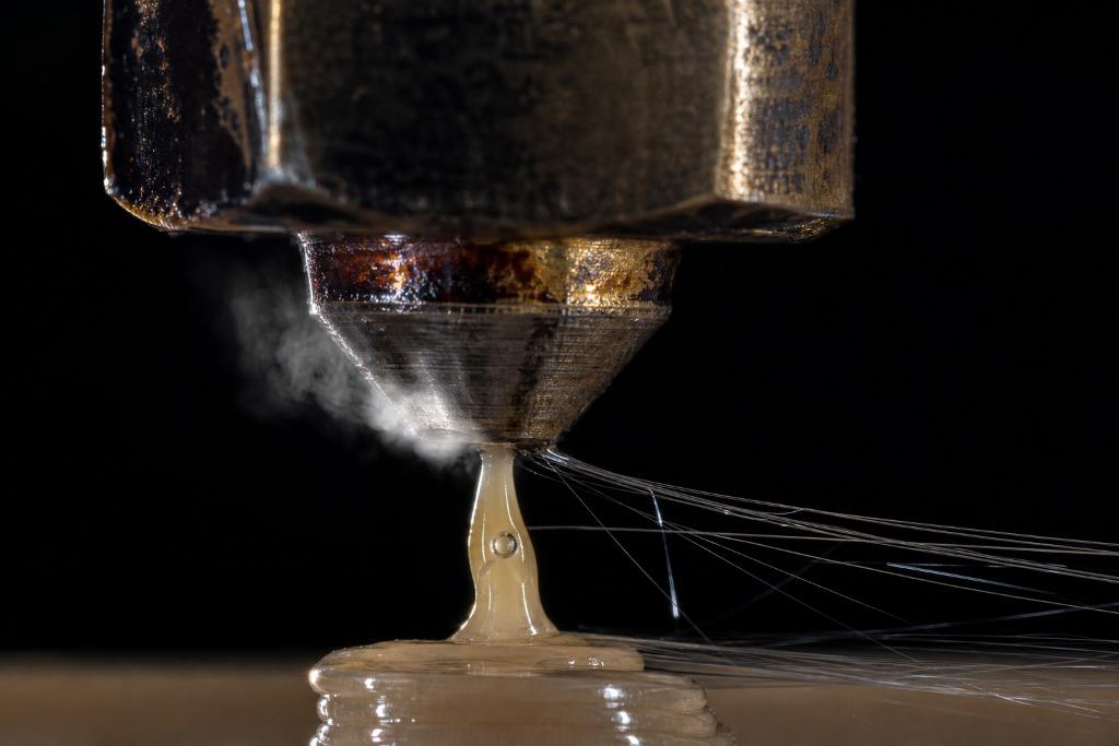 Close-up of a 3D printer nozzle extruding filament, with steam and thin threads visible, illustrating material settings.