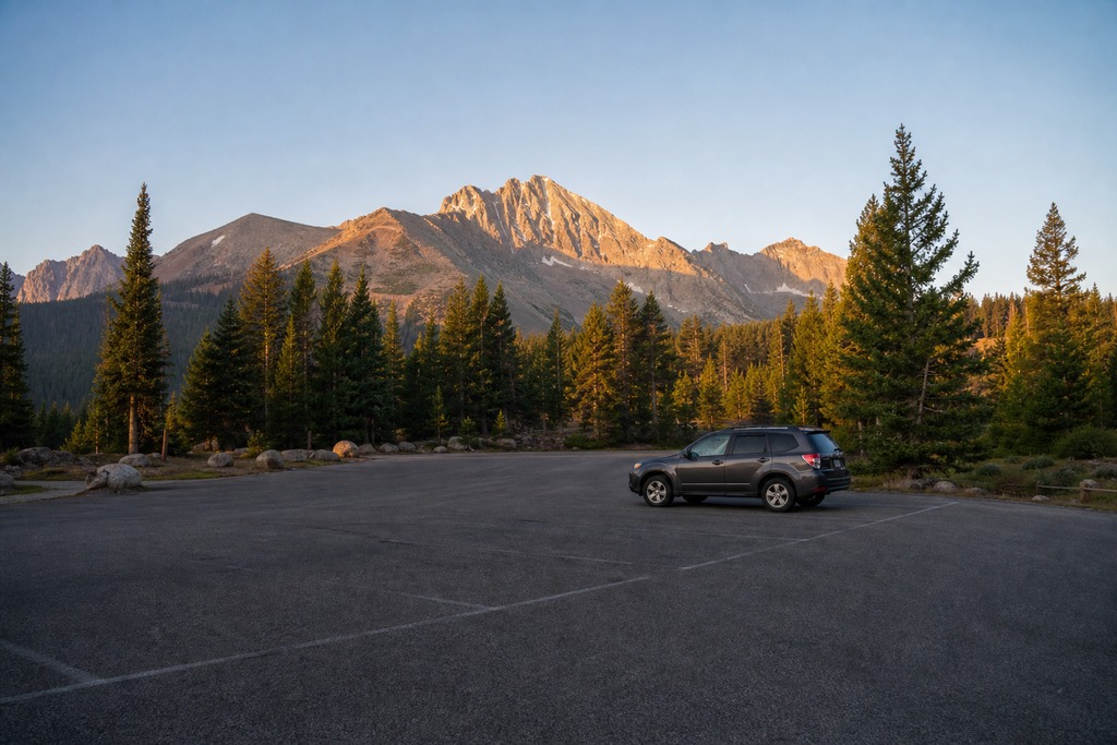 Parking lot with a lone vehicle surrounded by tall evergreen trees and majestic mountains at sunrise.